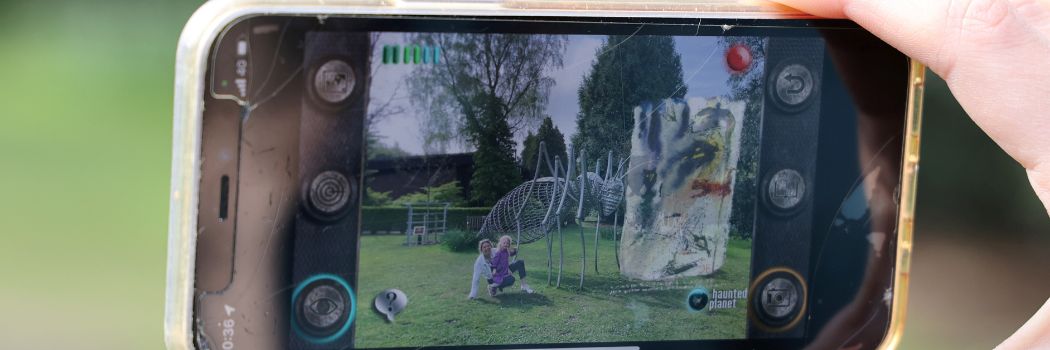 A mobile phone camera view of a woman and child on the grass next to a giant ant sculpture with AR artwork superimposed on the picture