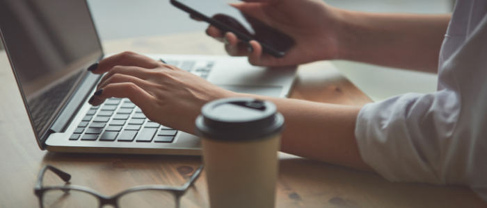 Person writing on laptop with phone, coffee and glasses