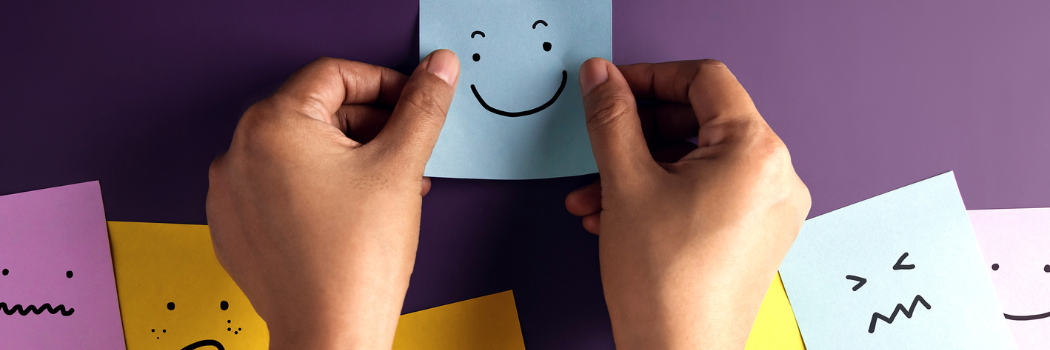 Person sticking a post it with a smile drawn on it onto a purple wall with other post it faces surrounding it