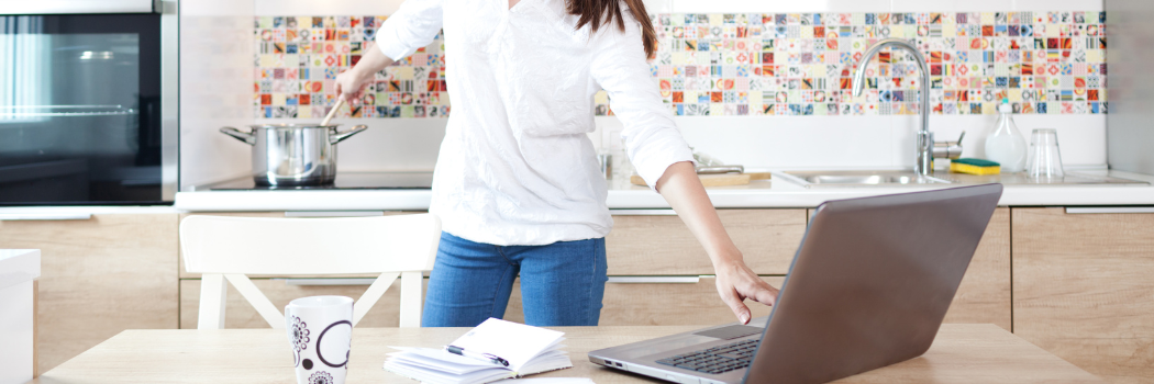 Woman in white shirt and blue jeans working on laptop and cooking at the same time