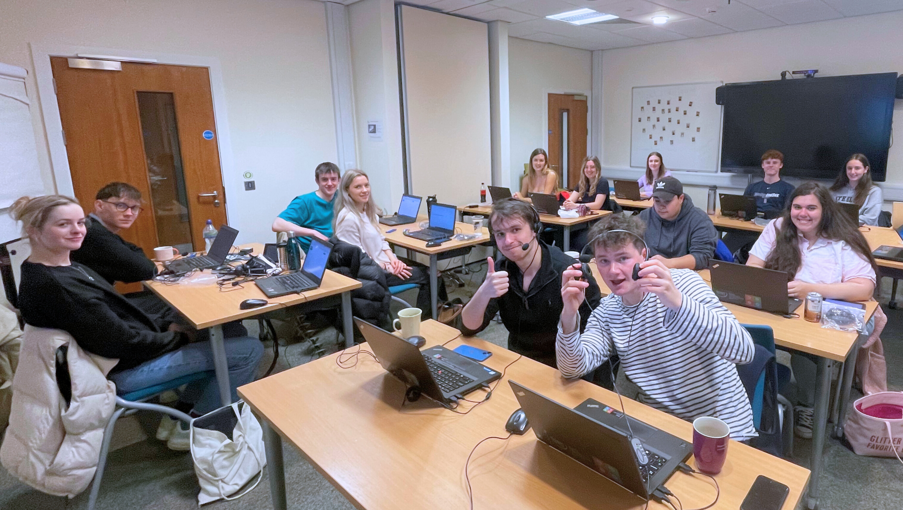 Students sitting in a room on laptops looking at the camera smiling