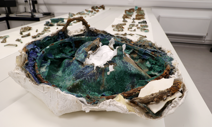 The crushed cauldron which was excavated as part of The Melsonby Hoard, with other artefacts from the hoard lined up on a table behind the cauldron.