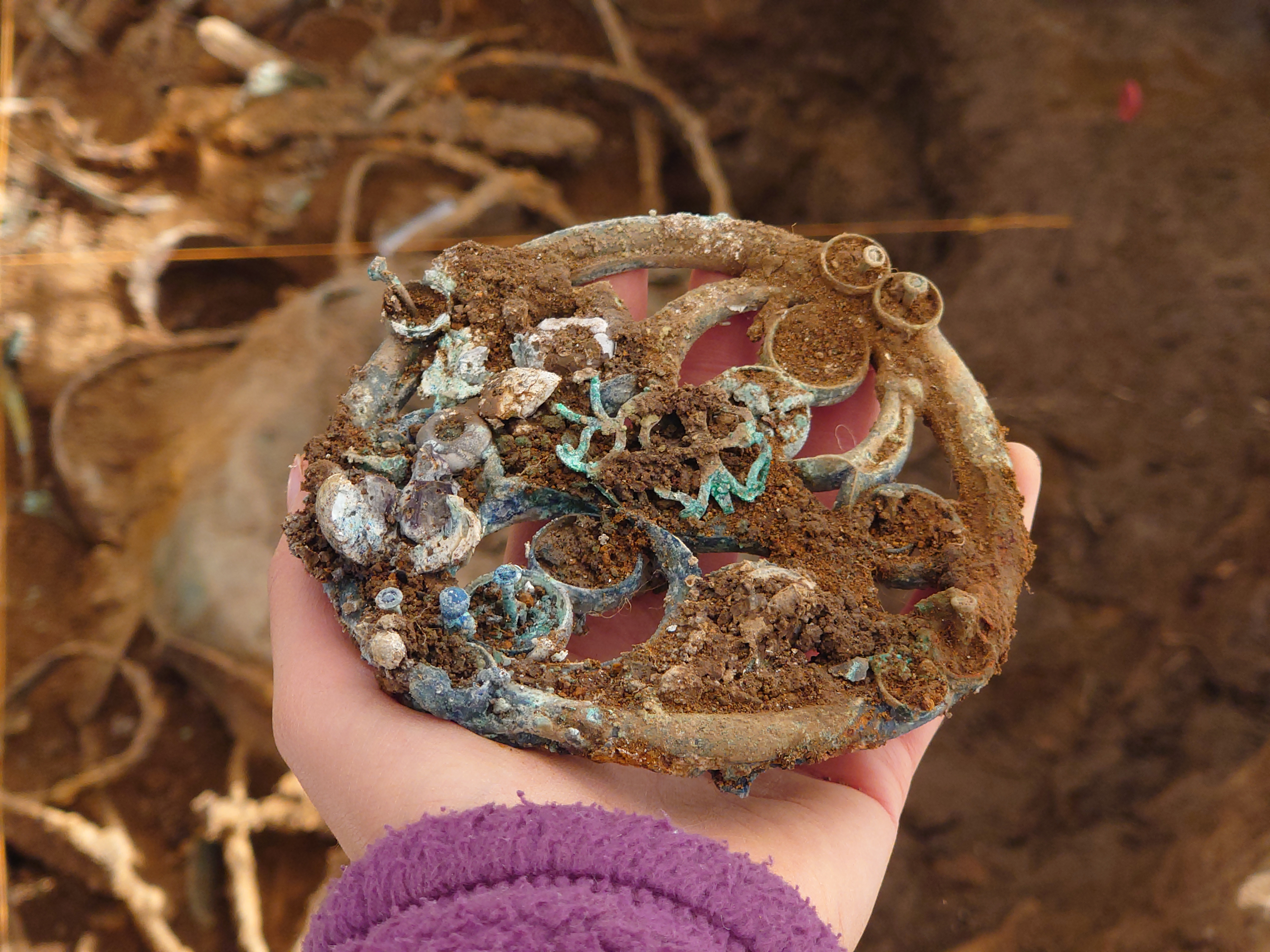 A close-up of a hand holding one of the artefacts from the Melsonby Hoard - a circular piece of embellished ironwork partially covered in soil. In the background, the excavation can be seen in progress.