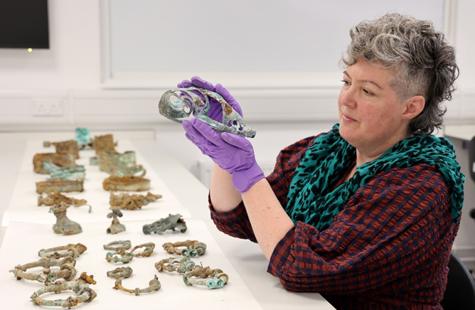 Dr Sophia Adams holding one of the artefacts from the Melsonby Hoard, with a number of other artefacts laid out on a table in front.