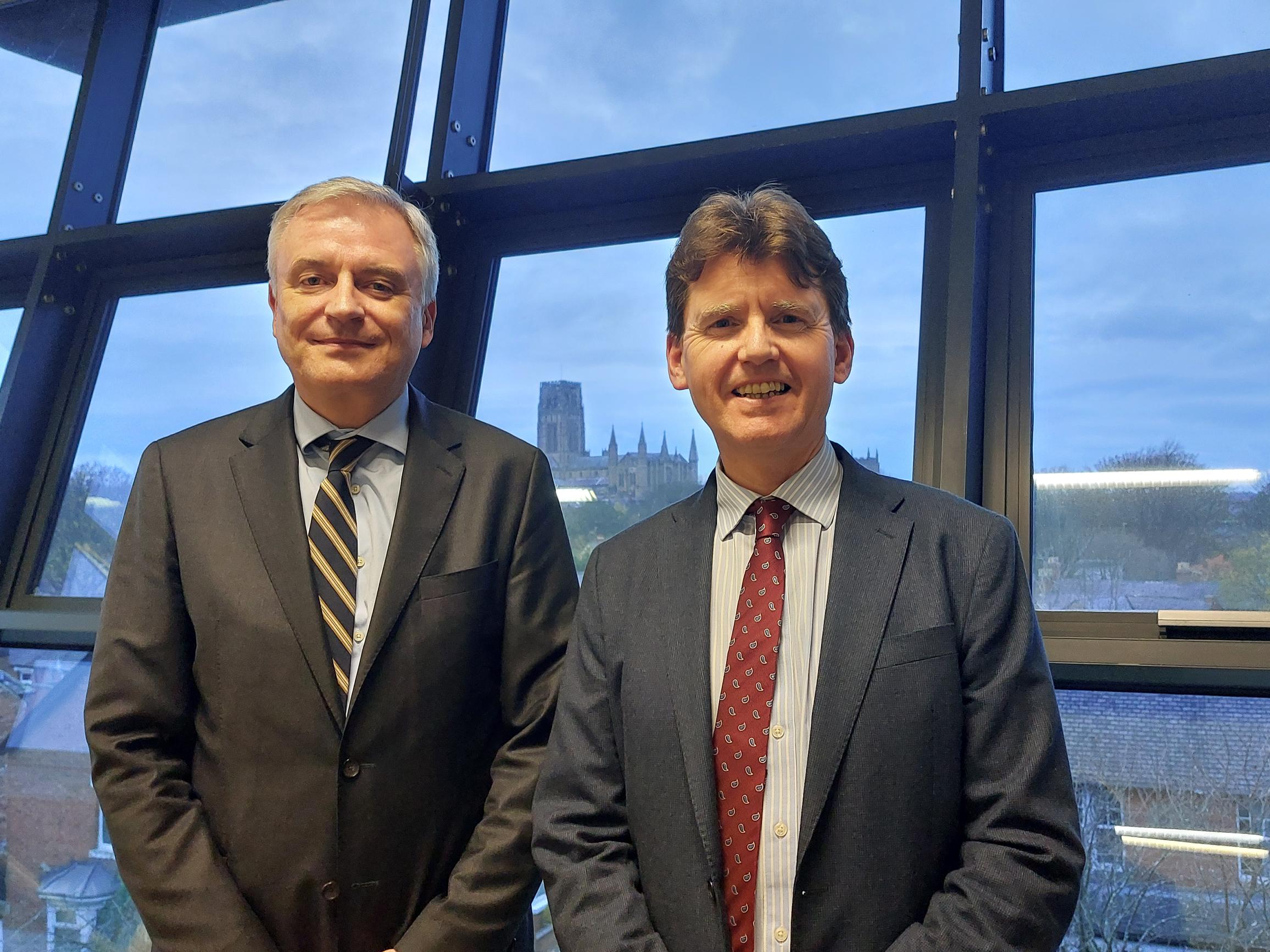 Two men wearing a suit and tie standing in front of a window