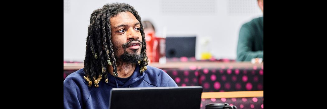 A black heritage male sitting in a lecture hall, behind a long desk, with a laptop, looking off screen