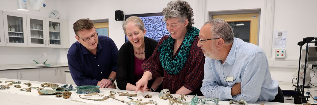A group of people looking and pointing as artefacts laid out on a lab bench.