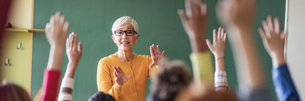 A female teacher talking to a classroom of pupils with their hands raised