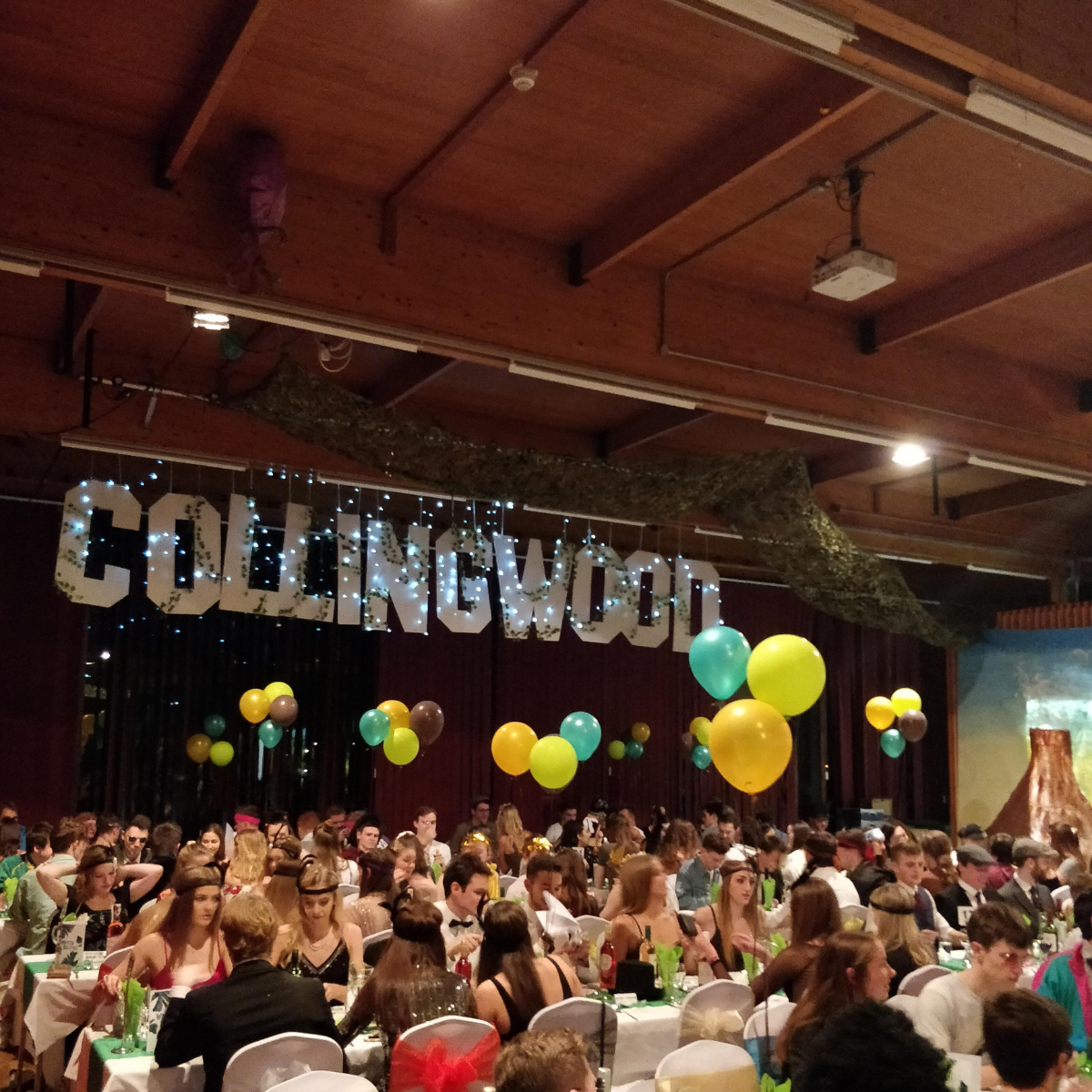 Students in formal wear at tables with balloons with Collingwood sign in background at formal dinner