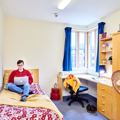 A picture of a male student siting on a bed in their accommodation with a computer