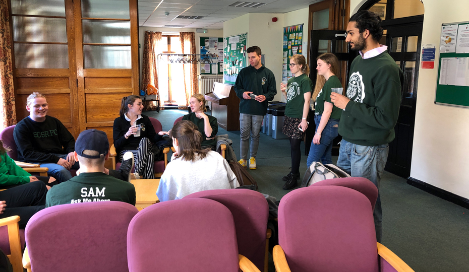 A group of students meeting in the Bailey Junior Common Room showing armchairs and posters.