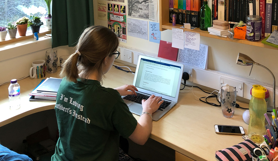 Female student sitting at a desk with a laptop with shelves above and a window to the side overlooking woodland.