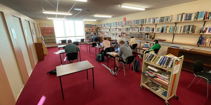 A group of students studying in the library space. There are books lining the walls and locked cupboards on one side containing older books