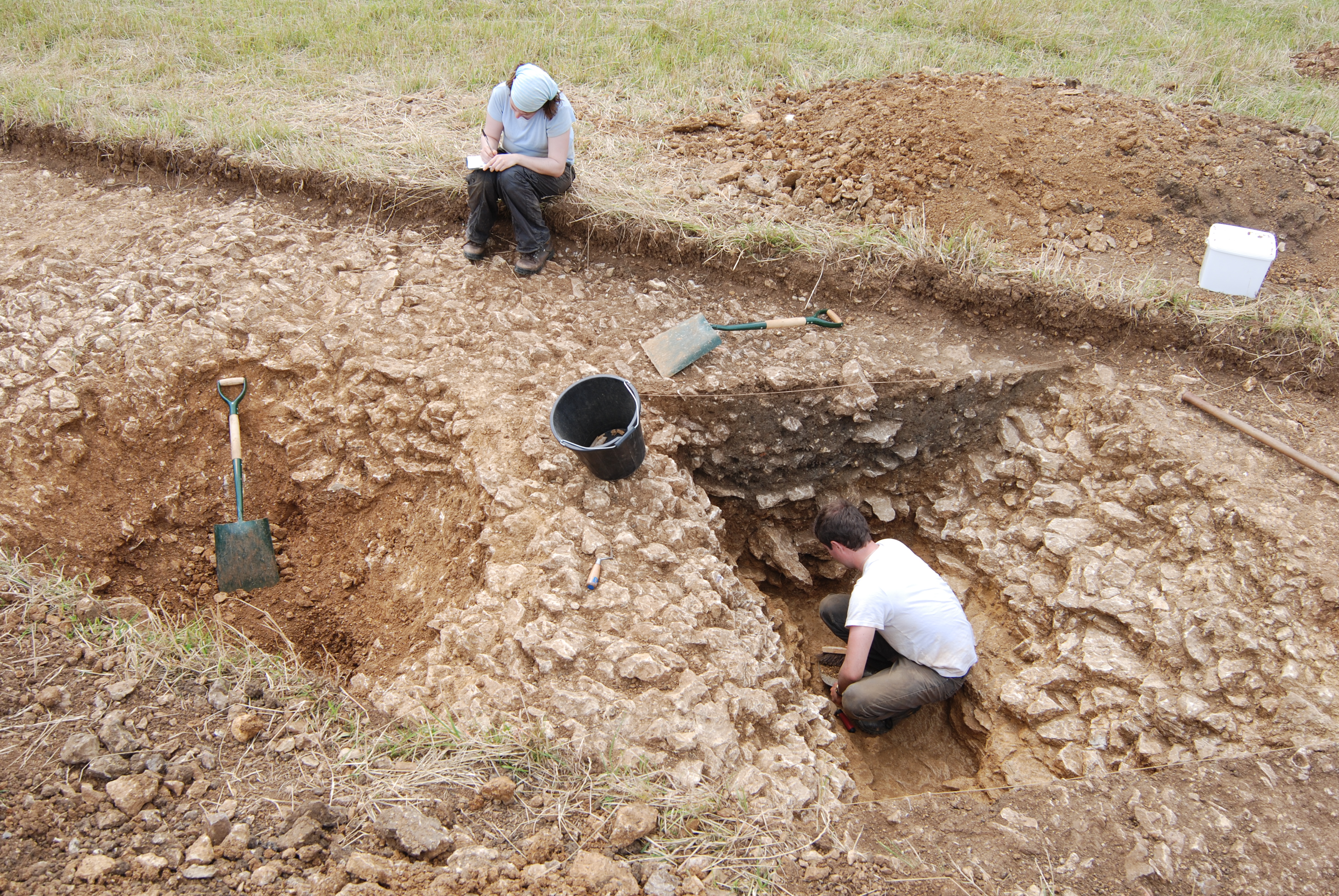 Two students on an archaeological dig with spades and a bucket