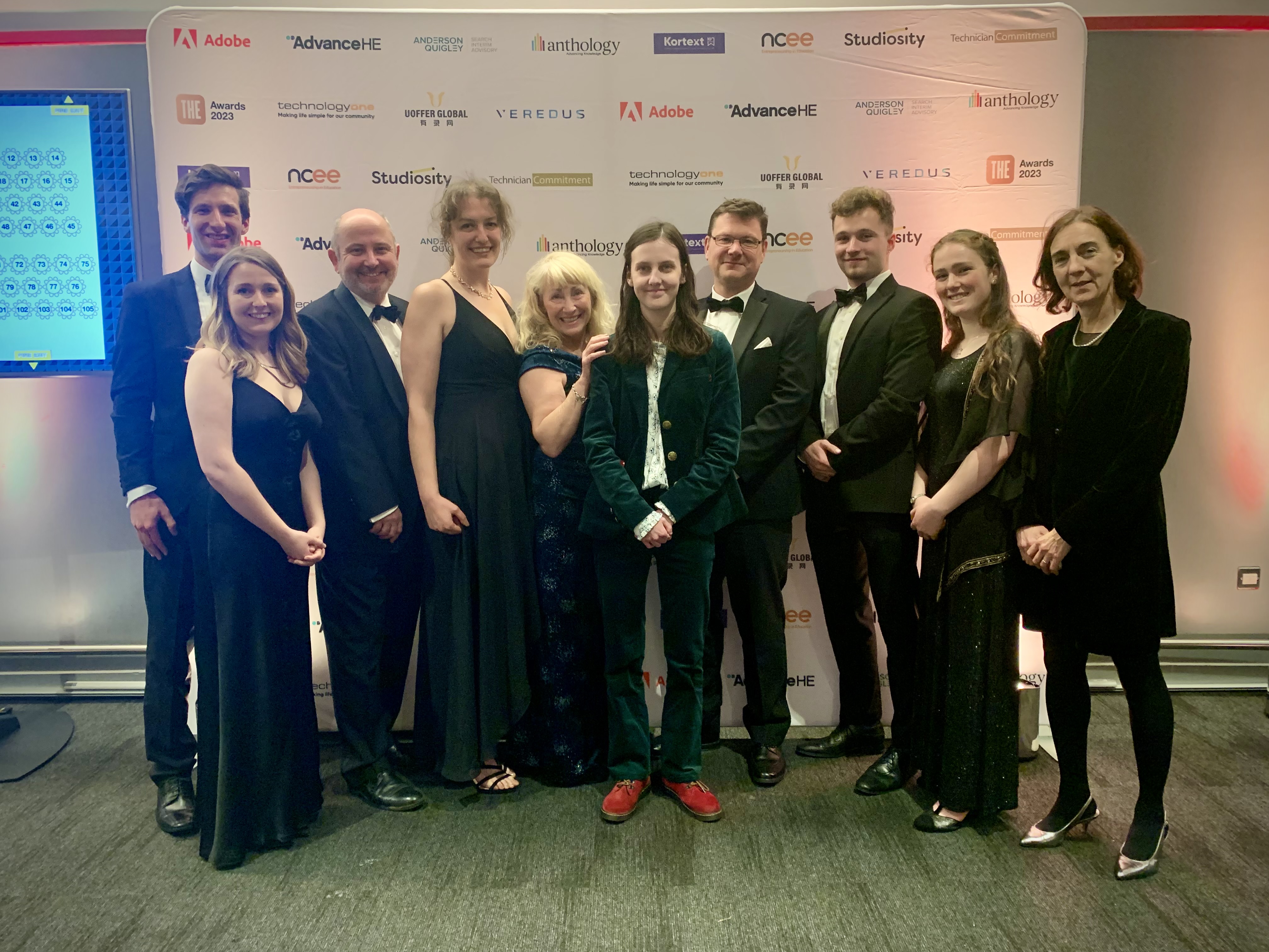 Group photo of 10 smiling people in black tie standing in front of a board featuring different company logos.
