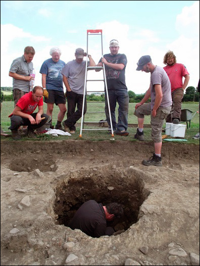 Group of archaeology students standing at the side of a trench watching an archaeologist digging in the bottom of a hole