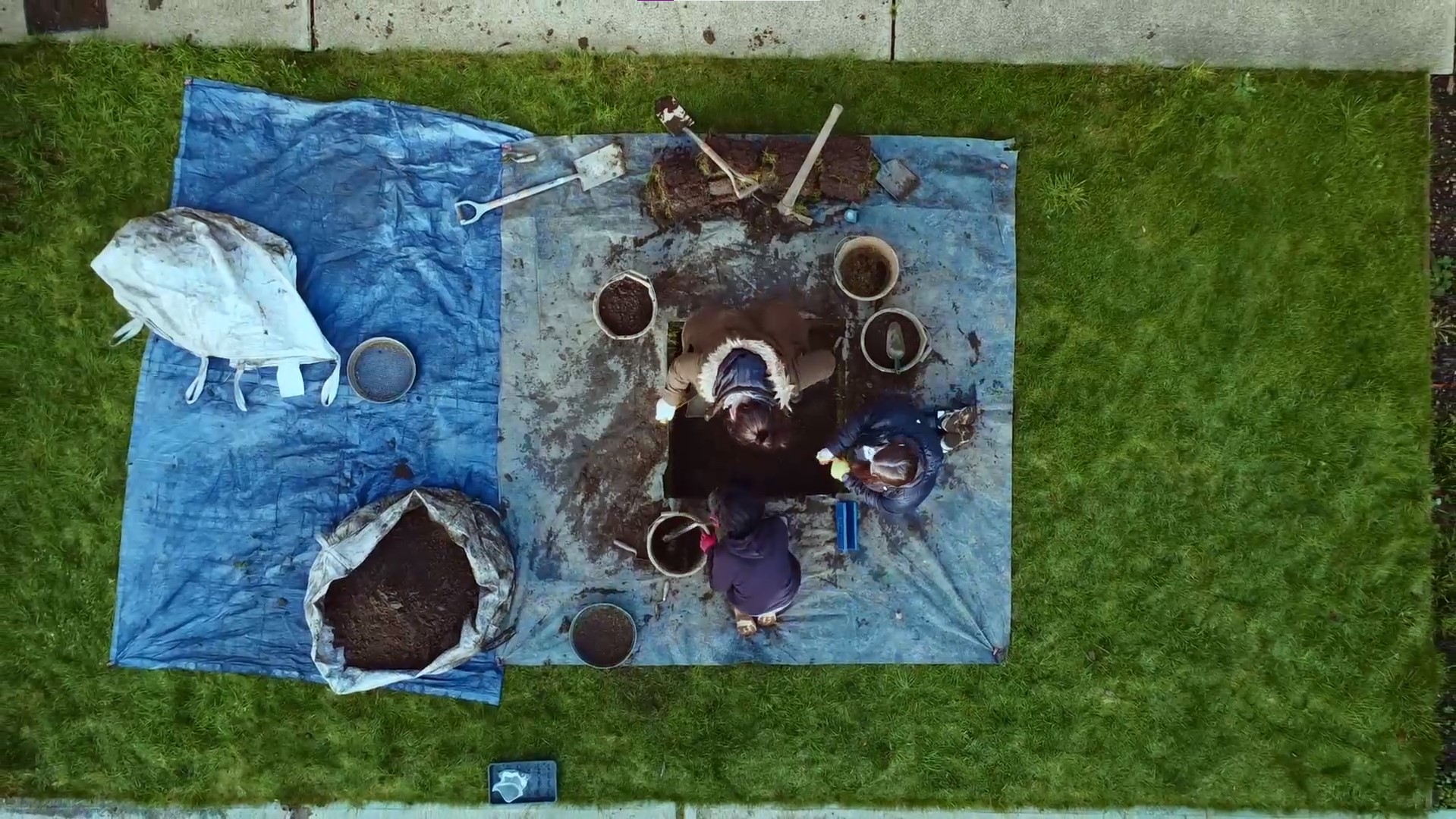 Aerial view of three warmly dressed people around a test pit dug in a grassy area. There are pots of soil, digging tools and blue sheeting around them. A paved surface is visible at the edge of the frame.