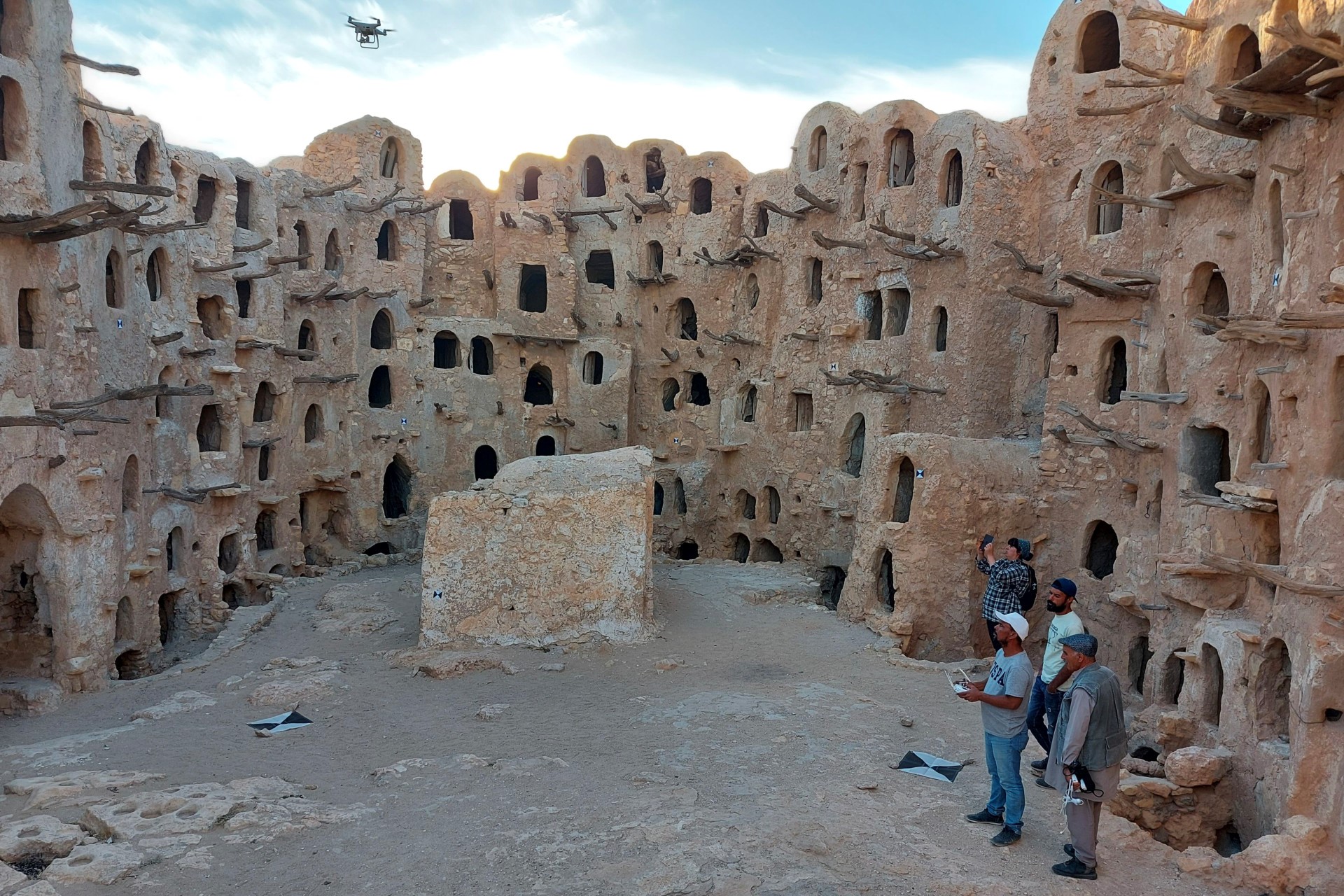 Archaeologists stand within the remains of stone buildings, and one of them controls a small drone flying above the buildings. They are located in Kasr Kabaw, Libya.
