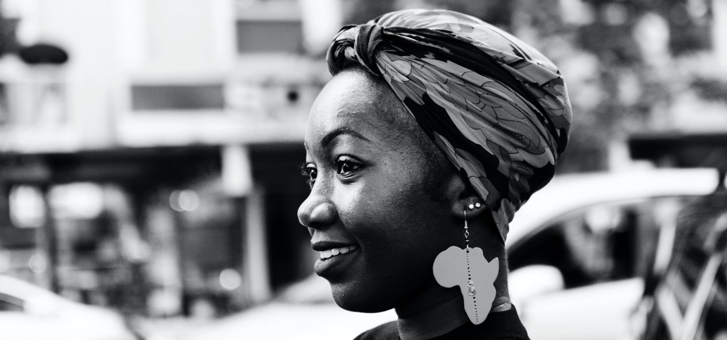  Monochrome photo of a Black woman standing side on to the camera, smiling and wearing an earring and headscarf. In the background is a busy city street.