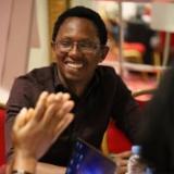 Candid photo of an early career researcher, David Muthegethi, wearing glasses, a dark brown shirt and smiling broadly, sitting in a table with a group who are out of frame. In the out-of-focus foreground are two hands that look to be mid-clap,