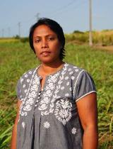 An early career researcher from Mauritius, Jayshree Mungur-Medhi, standing in a field in warm sunlight, wearing a grey and white embroidered shirt and looking at the camera. In the background are some electricity distribution poles.