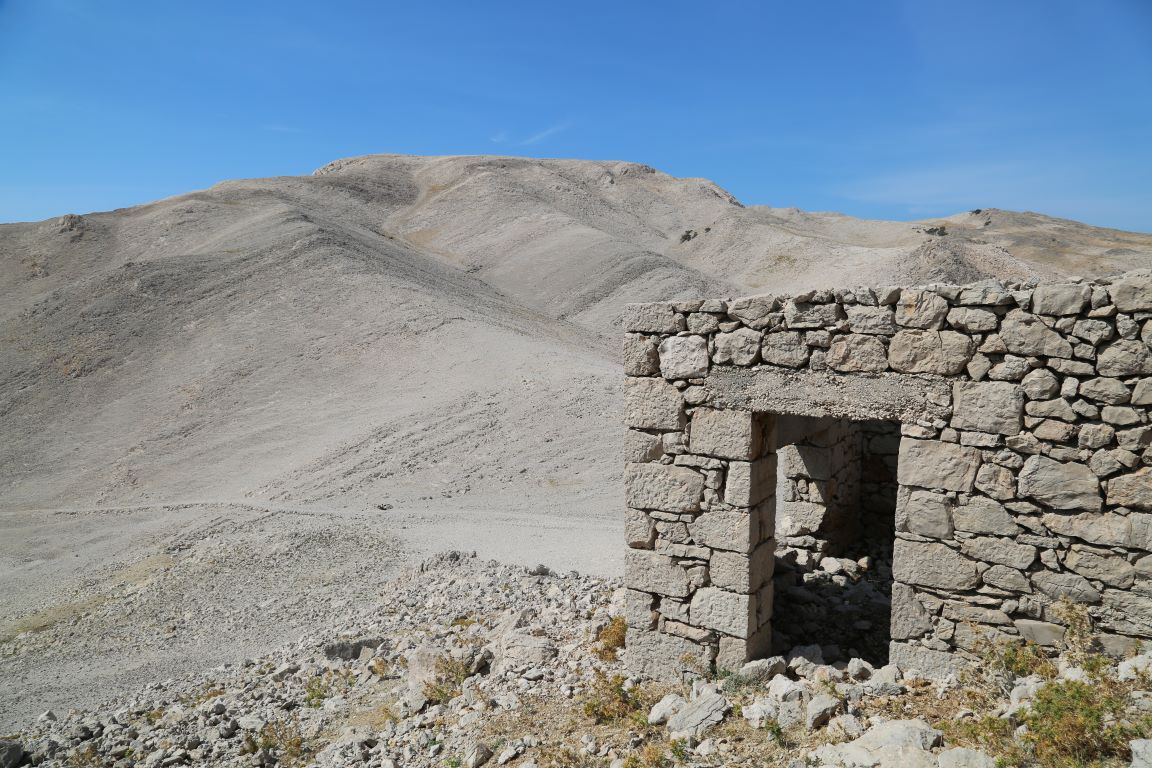 Remains of a stone building in front of rocky mountainous terrain and blue sky.