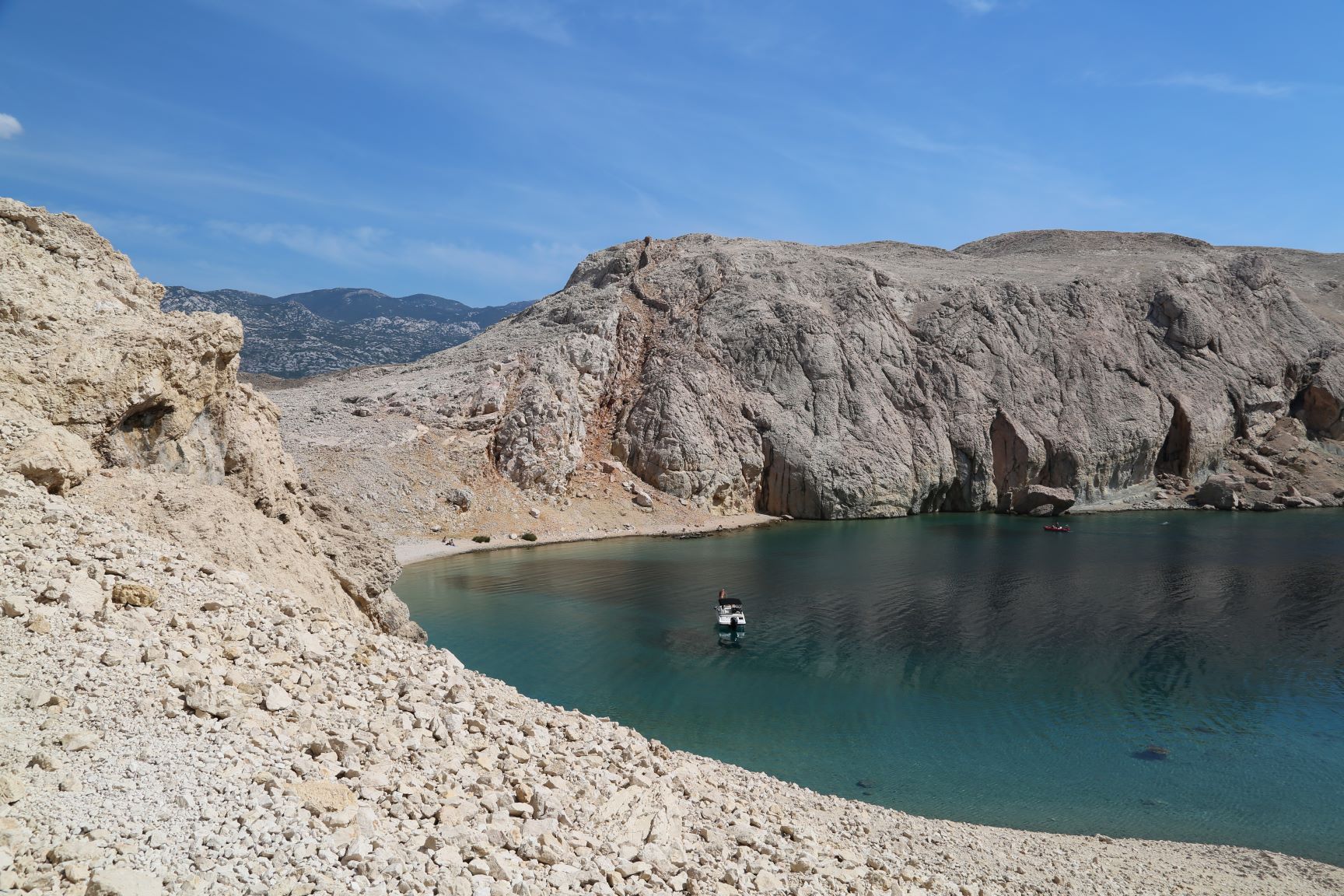 A small boat on a dark blue-green lake, bordered by rocky terrain. Distant mountains and blue sky are visible in the background.