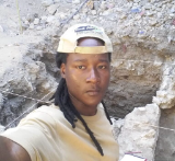 An early career researcher, Celso Simbine, wearing a beige backwards baseball cap and beige t-shirt, standing next to an excavated archaeological feature.