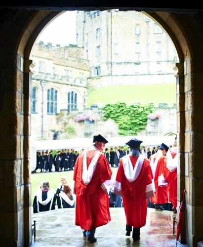 Two PhD graduates in red robes and hats walk out of a medieval doorway