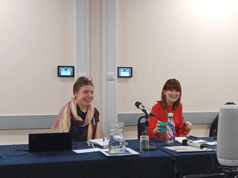 Photograph of two participants at the Organise! Organise! Organise! Conference, sitting behind a table. They are smiling at the camera. The table has microphones and papers, laptops and jugs and glasses of water.