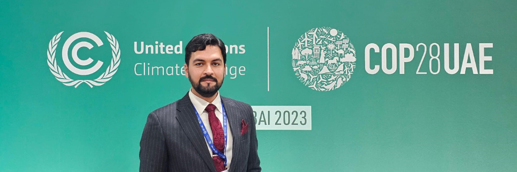 A man with a beard standing in front of a green sign displaying the COP28 logo.