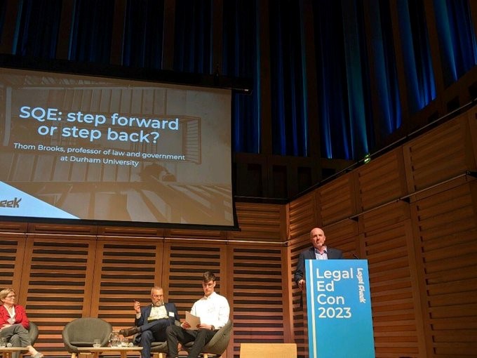 Thom is standing on a stage, behind a podium. There is a large powerpoint screen behind him.