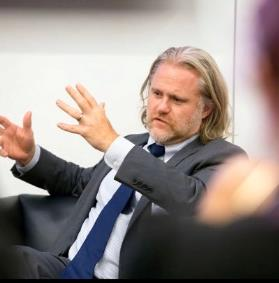 Charles T. Kotuby Jr sitting is a chair talking and gesturing with his hands. He is wearing a grey suit, white shirt and a navy tie. He has grey hair down to his shoulders and a short beard.