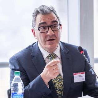 John Fellas sitting in front of a window. He is wearing glasses, a grey suit and you can see a microphone and a water bottle in the foreground