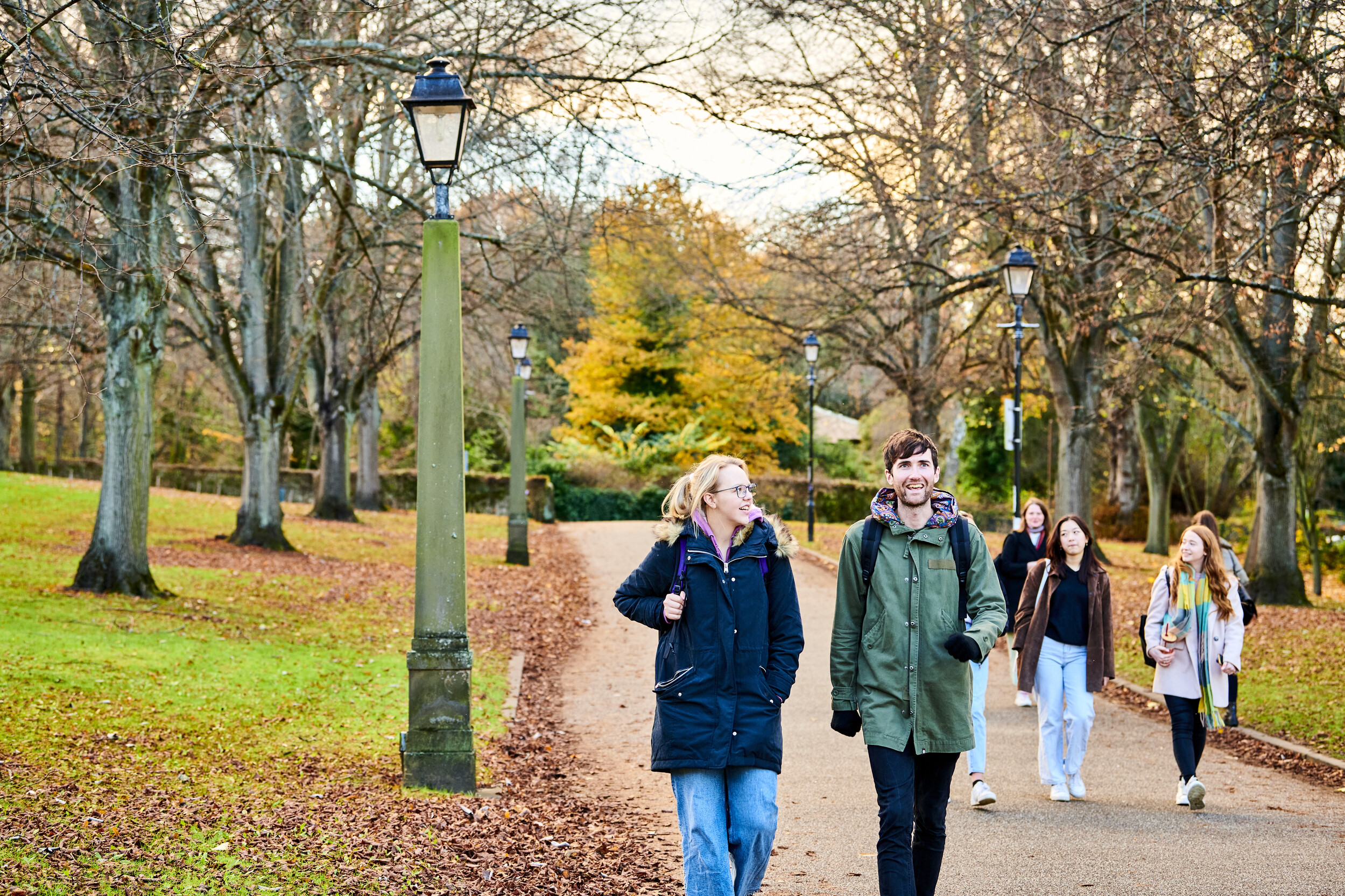 Male and female students walking along path with autumn trees in the background and leaves on the ground