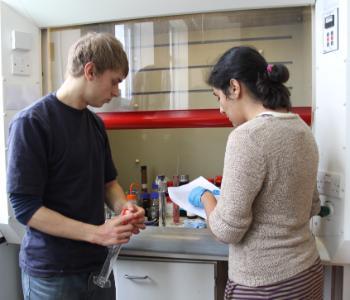 Male and female student check project notes, and the male holds a large test tube, whilst stood by a fume cupboard in a lab