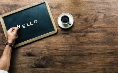 Closeup of a man sat at a table writing Hello on a chalkboard with a cup of coffee.