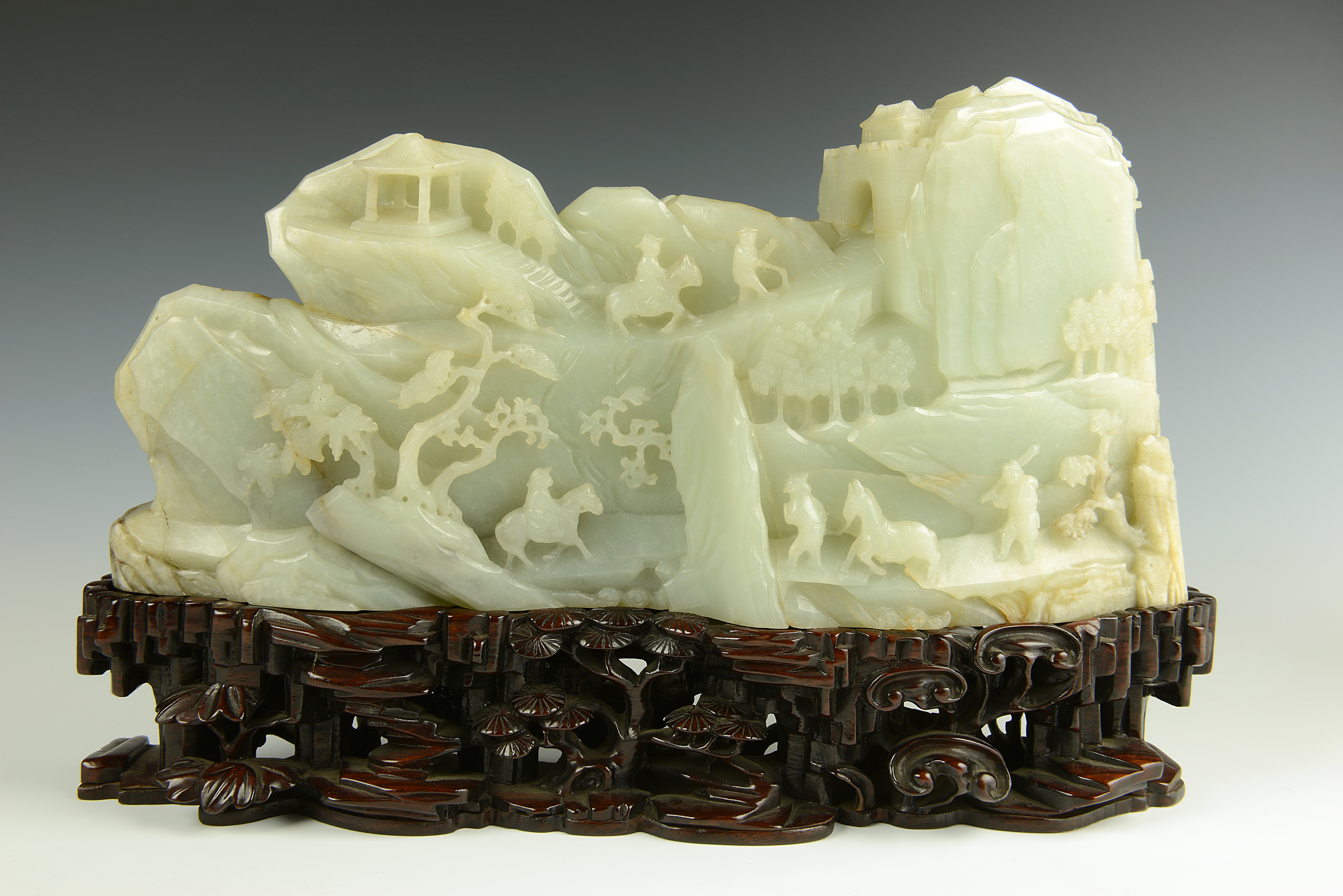 A carving in pale green jade of a mountain scene with trees and two human figures travelling along the side of the mountain, one on horseback and the other on foot carrying baskets over his shoulder. The carving stands on a carved wooden stand.
