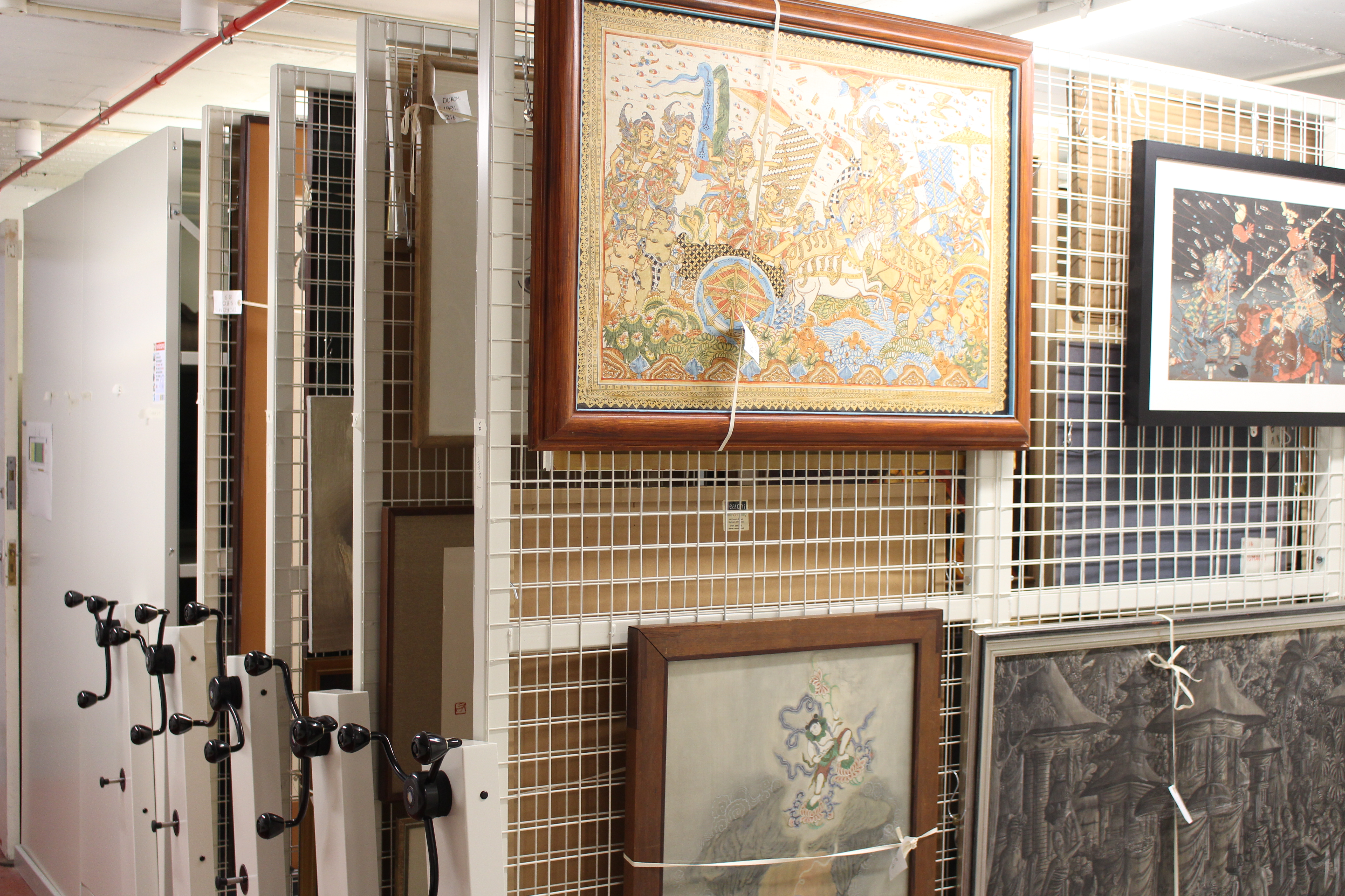 paintings hanging on a series of vertical mesh racks with other cupboards visible behind