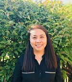 Headshot of Mel Earnshaw, she is brown skinned and wearing a black shirt and is standing on front of a leafy hedge.