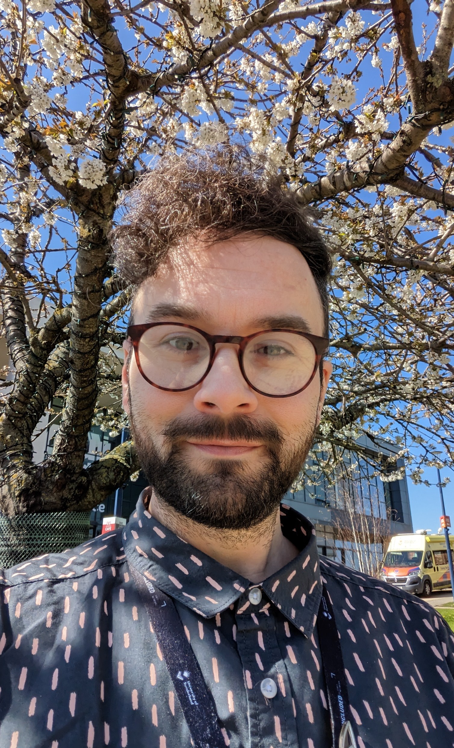 Andrew is light skinned with brown hair and a brown beard. Andrew is wearing glasses and a Navy and pink shirt. In the background are blue skies and a blossom tree