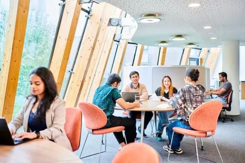 Groups of postgraduate students sit in a modern, airy study space. It has high windows and ceilings with lots of pale wooden panels and colourful furnishings. The students are sitting with laptops in front of them and are chatting with one another. They look relaxed and happy