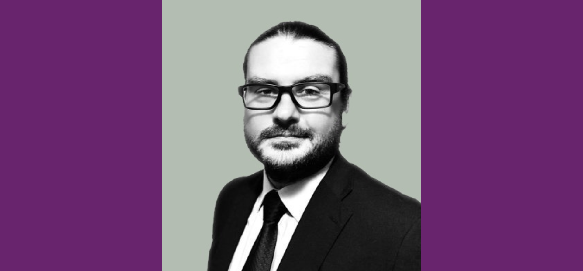 A black and white photo of a man wearing a suit, with a beard, glasses, and long hair tied back