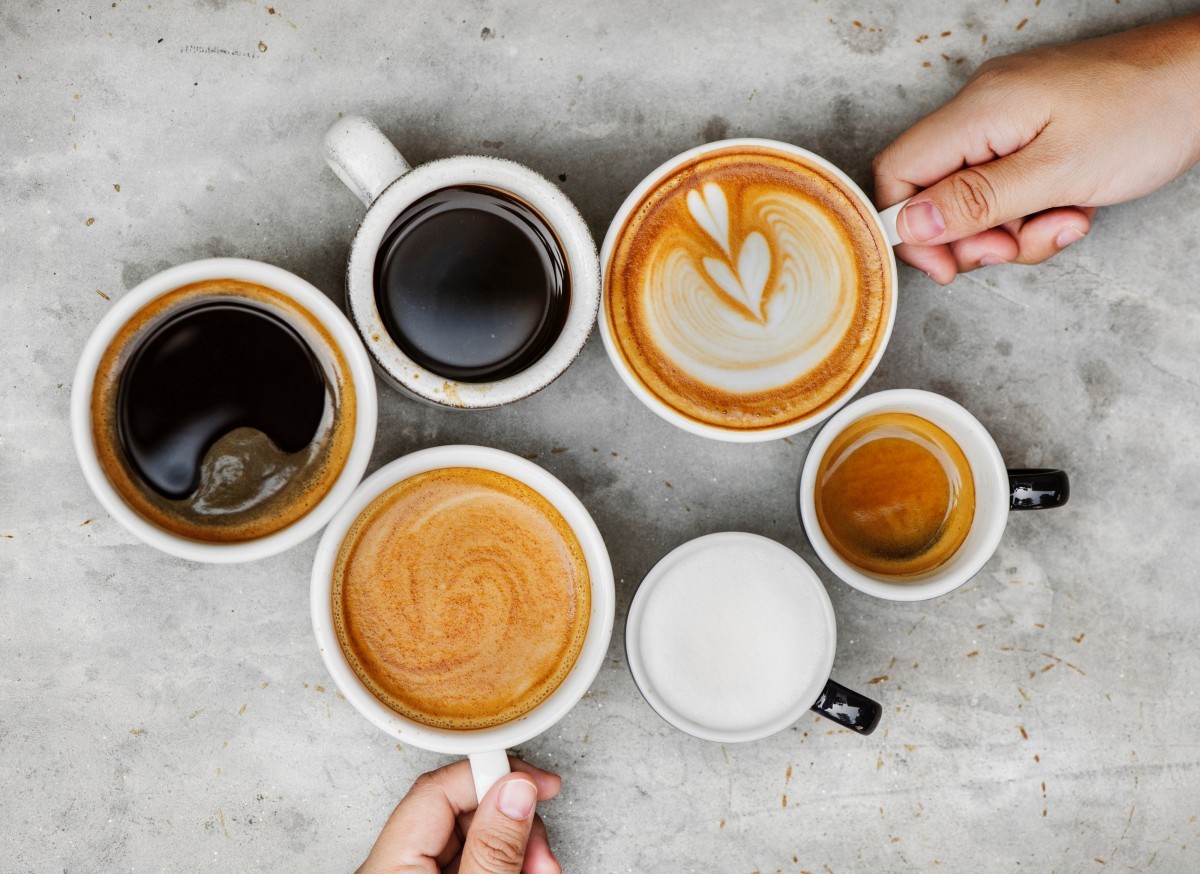 Birds eye view of 6 coffee cups filled with different types of coffee and a hand on 2 of the handles.