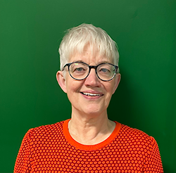 A headshot of a smiling white woman with glasses standing in front of a green wall