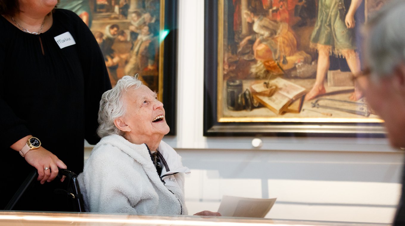 An older person in a wheelchair looks up at the art on the walls of a gallery and smiles.