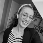 Black and white photo of Georgia Punton smiling at the camera (young white girl with long curly hair in a ponytail wearing a headband, glasses, striped shirt and cardigan)