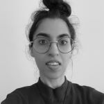 Black and white photo of Veenu Gupta (young Asian woman with medium-length dark hair in a bun wearing glasses and a high neck jumper)