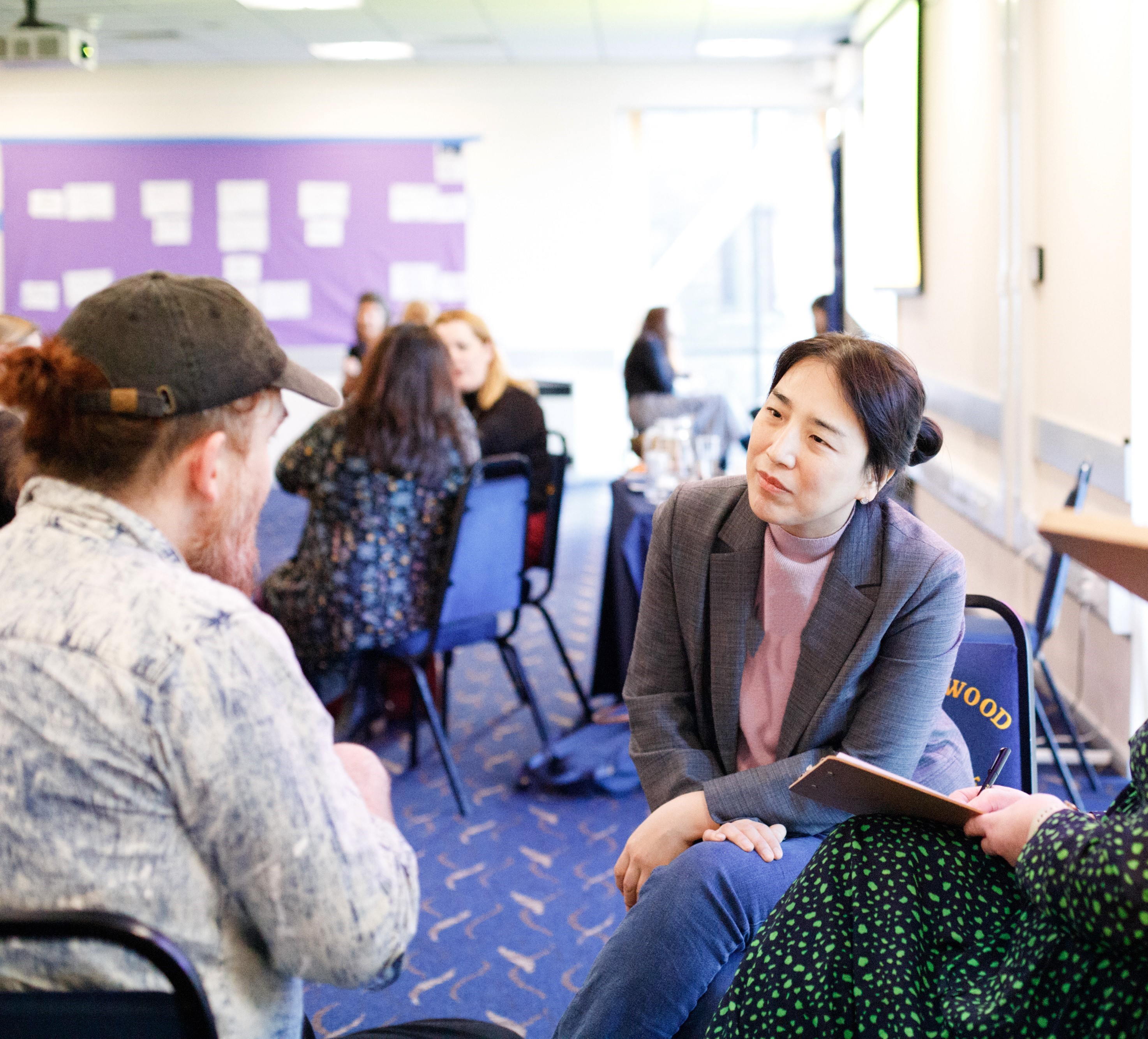 A group of three people sat in discussion - on the left, the back of a white individual with a beige shirt and khaki baseball cap; in the middle, an asian woman with dark hair in a bun, wearing a pink t-shirt and grey blazer; on the right, the legs of a white woman wearing a full-length dark green dress