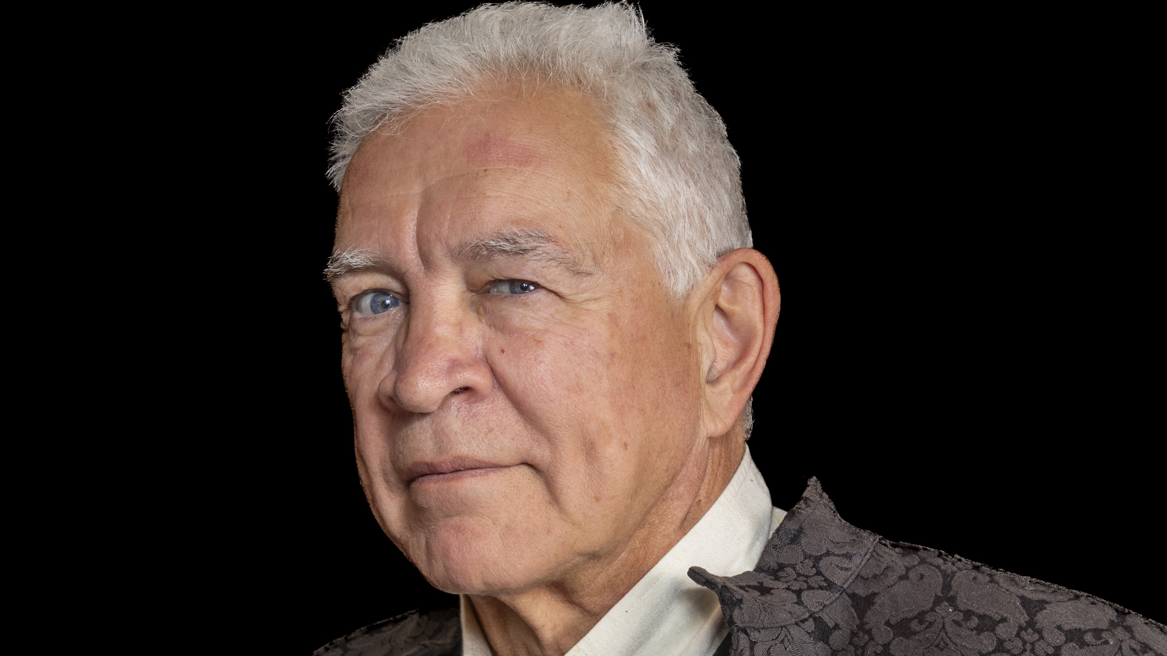 Older white man with short white hair staring at the camera in a white shirt and a grey jacket.